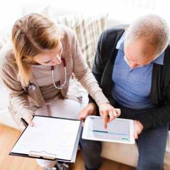 Health visitor and a senior man during home visit. A female nurse or a doctor showing test results on a tablet. High angle view.